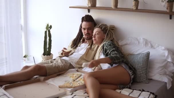 Casal encantador com dreadlocks ter uma manhã preguiçosa comer pizza entrega e assistir TV enquanto deitado na cama. Menina loira usando controle remoto, escolhendo os canais — Vídeo de Stock