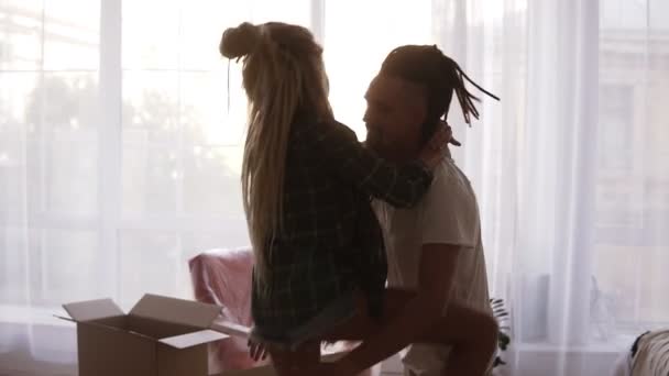 Hipsteři pohybující se pár jsou nadšení, že se stěhují do nového bytu, zvedne ji do náručí a oni se točí kolem - hromady lepenkových krabic v pozadí — Stock video