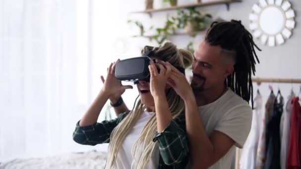 Technologia, rozszerzona rzeczywistość, koncept gier, rozrywki i ludzi - szczęśliwa para - dziewczyna próbująca wirtualnych słuchawek lub okularów 3D grających w domu. Jej chłopak stoi za nią i wspiera — Wideo stockowe