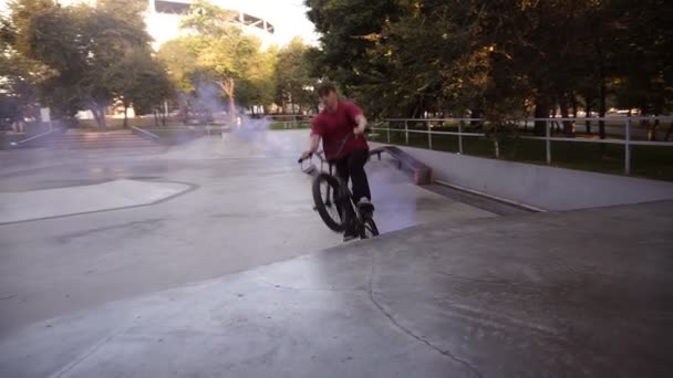 Молодий кавказький гонщик, який практикує у порожньому скейт-парку, їде на пандусі, а блакитна димова бомба горить на велосипедах заднім колесом. Чоловік практикує трюки, їде на велосипеді і розважається. Повільний рух — стокове відео
