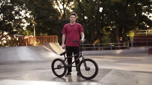 Um jovem com uma bicicleta BMX fica ao lado de sua bicicleta com rampa, parque de skate no fundo. Retrato do cavaleiro BMX. Cultura de rua. Infláveis de lente no fundo. Steadicam baleado. Vista frontal — Vídeo de Stock