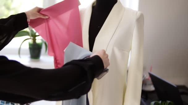 Un tailleur créatif non identifié essaie différents échantillons de tissu de couleurs sur une veste blanche sur un mannequin dans son atelier pour coudre des vêtements. La créatrice de mode prépare un nouveau modèle de vêtements qu'elle produit — Video