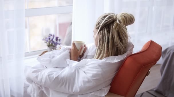 Chica rubia con rastas largas mira por la ventana en acogedor piso. Mujer sentada cerca de la gran ventana sosteniendo jarra y bebiendo. La señora se sienta en una silla cubierta de manta blanca en sus pensamientos. Atrás. — Vídeos de Stock