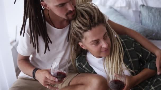 Una pareja moderna enamorada de rastas bebiendo vino tinto de copas de vino. Disfrutando del fin de semana libre juntos acostados en la cama abrazando. De ocio familiar. Movimiento lento — Vídeos de Stock