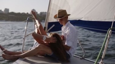 Genç romantik çift, yatların üzerinde oturmuş kucaklaşıyorlar. Uzun saçlı esmer kız erkek arkadaşının dizlerinin üzerinde yatıyor. Beyaz tişörtlü ve şapkalı bir adam onu okşuyor.