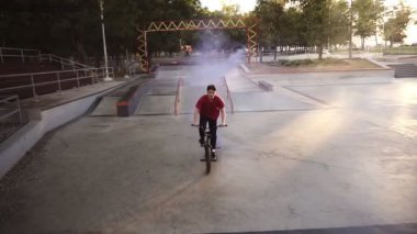 Mavi duman bombası arka tekerleklerde yanarken, kaykay parkında pratik yapan genç beyaz bmx binicisi. Adam numara yapıyor, bisiklete biniyor ve eğleniyor. Yavaş çekim