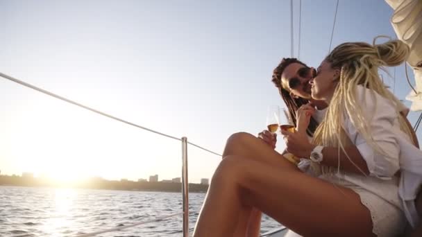 一对穿着白色衣服、戴着太阳镜的时髦夫妇坐在游艇边缘，微笑着，与冠军眼镜叮当作响。 爱恋的夫妻在游艇上共度时光 — 图库视频影像