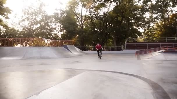 Cool bmx motociclista em t-shirt vermelha e calças esportivas pretas pulando, fazendo 360 graus virar truque no belo dia de verão no parque de skate. Movimento lento — Vídeo de Stock
