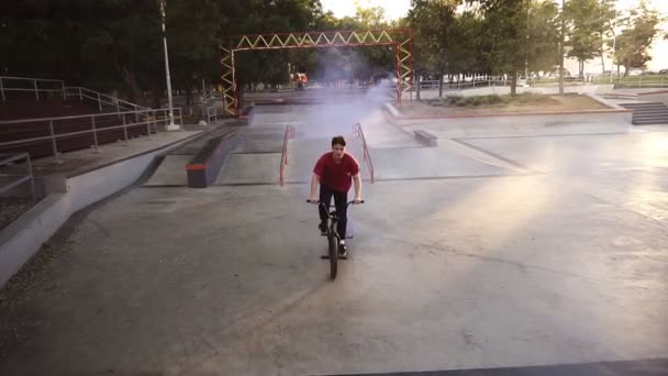 Молодой кавказский гонщик bmx практикуется в пустом скейт-парке, катается на рампе, в то время как голубая дымовая бомба горит на велосипедах заднего колеса. Человек практикует трюки, катается на велосипеде и веселится. Медленное движение — стоковое видео
