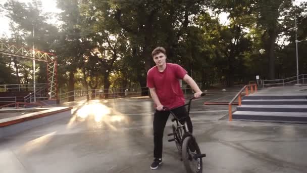 Молодой человек наслаждается велосипедом BMX, катается вокруг, стоит на педалях и останавливается от переднего вида, позируя перед камерой на велосипеде. Bmx спортсмен, занимающийся в скейт-парке — стоковое видео