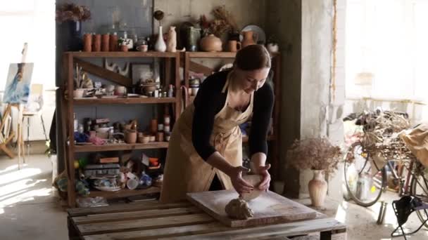 Retrato de una alfarera vestida con delantal beige que pone una pieza de arcilla en la encimera y luego comienza a amasarla con las manos. Productos de cerámica en el estante detrás — Vídeo de stock