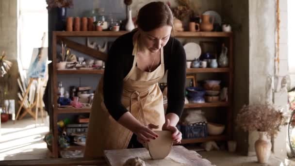 女性陶工身穿米黄色围裙，在工作台上轻轻揉捏粘土片，用手工作的前景。架子上的陶器制品，镜头照明弹 — 图库视频影像