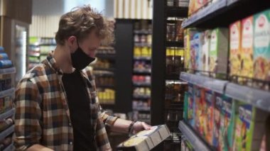 Salgın sırasında süpermarkette erkek müşteri. Makarna paketinin etiketini okuyan adam. Raflar ve bölüm sıraları arka planda. Siyah koruyucu maske ve eldiven takmış.