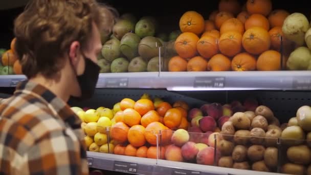 Ekose gömlekli genç adam, solunum cihazı takıyor, karantina sırasında markette plastik poşetli meyve alışverişi yapan siyah mendil maskesi giyiyor. — Stok video