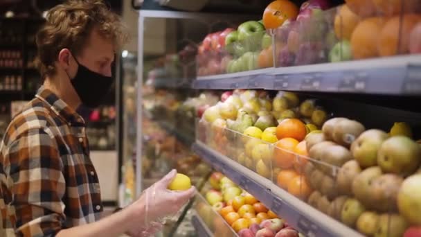 Молодой человек в клетчатой рубашке, в черной маске из ткани и пластиковых прозрачных перчатках, покупает фрукты, выбирает лимоны в супермаркете во время карантинного пандемического ковируса-19. Side — стоковое видео
