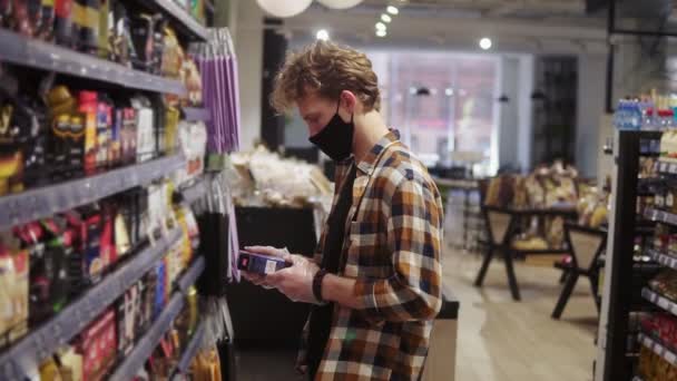 Πελάτης στο σούπερ μάρκετ κατά τη διάρκεια πανδημίας. Ο άνθρωπος διαβάζει την ετικέτα στο πακέτο του καφέ. Ράφια στο παρασκήνιο. Φορώντας μαύρη προστατευτική μάσκα — Αρχείο Βίντεο