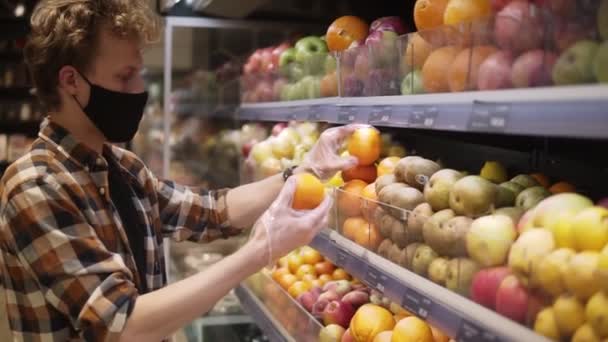 Mladý muž v kostkované košili, s hedvábnou černou maskou a průhlednými plastovými rukavicemi, nakupující ovoce v supermarketu během karanténního pandemického covid-19 koronaviru. Boční pohled — Stock video