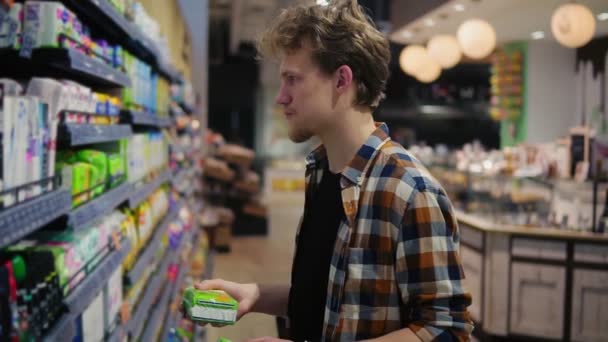 Jovem caucasiano segurando almofadas femininas no shopping e lendo o rótulo, risonho. Conceito de compras e escolhas. Vista lateral. Movimento lento — Vídeo de Stock