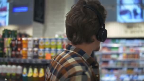В супермаркете: белый парень в наушниках ходит по товарной секции магазина, просматривает. Следую за кадром сзади. Медленное движение. Закрыть — стоковое видео