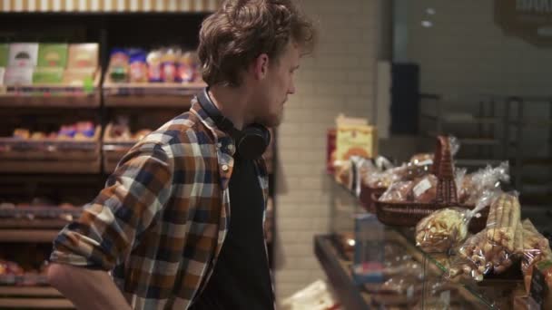 Boynunda kulaklık olan genç adam lezzetli kekleri seçiyor, modern süpermarket fırınında cam vitrinde hamur işlerini seçiyor. Yan görünüm — Stok video