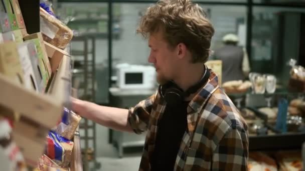 O homem de camisa xadrez escolhe um pão fresco no supermercado. Um jovem a tirar dois tostões de pão da prateleira coberto de sacos. Compras na mercearia — Vídeo de Stock