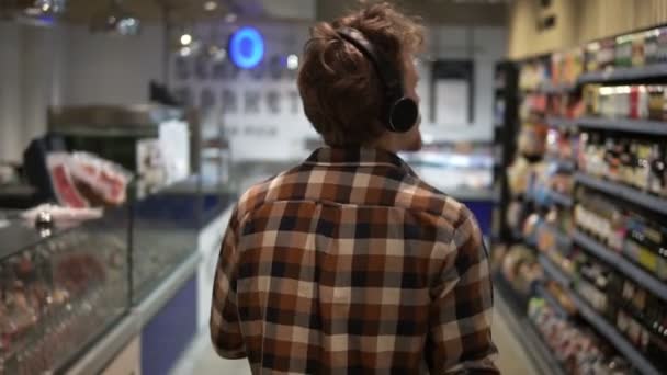 Snygg kaukasier kille med hörlurar promenader genom varor avsnitt av butiken, bär rutig skjorta - lyssna på bra musik.. Följer bakgrundsbild. Långsamma rörelser — Stockvideo