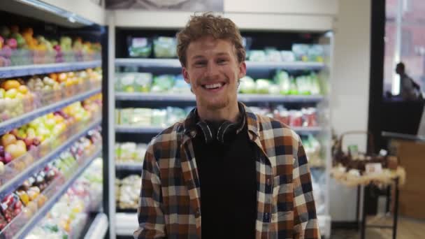 接近那个年轻英俊的高加索人，带着耳机在超市里购物，对着摄像机开心地笑着。肖像画 — 图库视频影像