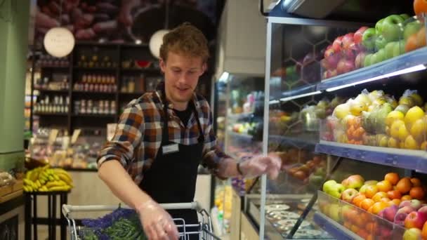 En el supermercado: Guapo empleado de bolsa con delantal negro, organizando frutas y verduras orgánicas. Añadir manzanas rojas frescas en el estante de la tienda. Movimiento lento — Vídeo de stock