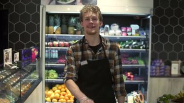 Kameranın önünde önlüklü genç beyaz bir dükkan işçisinin portre resmi. Grossery Supermarket 'te tezgah iterken neşeyle gülümsüyor.