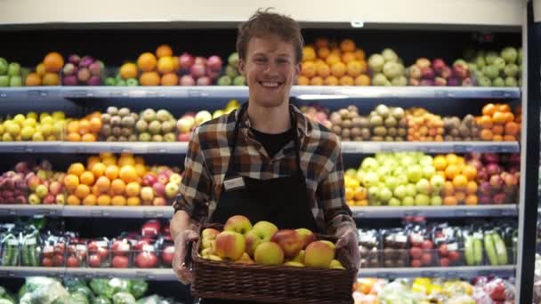 Retrato del joven trabajador caucásico guapo en el delantal parado frente a la cámara y sonriendo alegremente mientras sostiene la caja de manzanas en el supermercado grossero. Frutas y hortalizas — Vídeo de stock