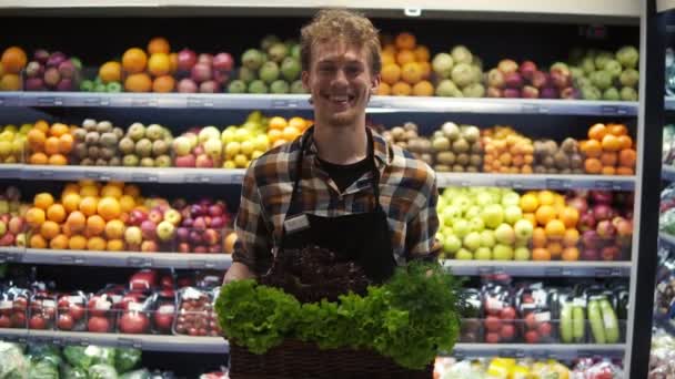 カメラの前に立ったエプロン姿の若いハンサムな白人店員の肖像画を撮影し、興行収入スーパーでボックスの色の緑を保持しながら楽しそうに微笑んだ。果物・野菜 — ストック動画