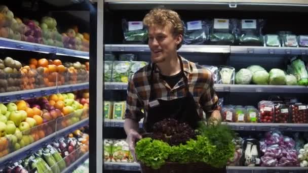 Szczęśliwy sprzedawca twarzy człowiek w supermarkecie przechadzając się po przejściu warzywa z pudełkiem świeżych zieleni do zorganizowania. Pracownik kaukaski w lokalnym supermarkecie — Wideo stockowe