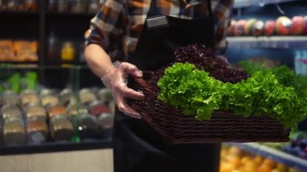 Vendedor homem em avental no supermercado andando por vegetais corredor com caixa de verdes frescos para organizar. Trabalhador caucasiano em supermercado local segurando caixa de verduras. Fechar — Vídeo de Stock