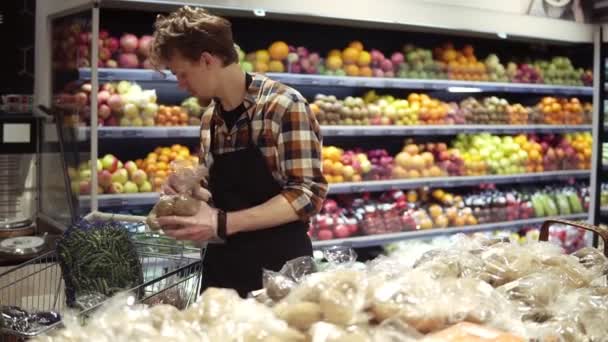 Ocupado trabajador joven caucásico que organiza verduras en los estantes en la tienda de comestibles. Chico en delantal llenando estante de almacenamiento con papas orgánicas saludables en el supermercado — Vídeos de Stock