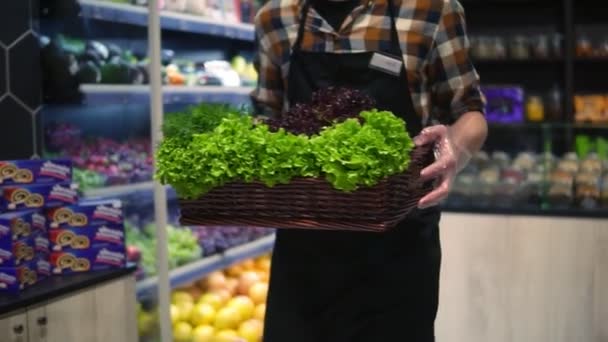 Venditore uomo in grembiule nel supermercato a piedi dalla navata verdure con scatola di verdure fresche da organizzare. Lavoratrice caucasica in un supermercato locale con una scatola di verdure. Chiudete. Rallentatore — Video Stock