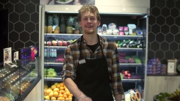 Portretfoto van de jonge knappe blanke winkelier in het schort voor de camera en vrolijk lachend terwijl hij de kar duwde in de supermarkt — Stockvideo