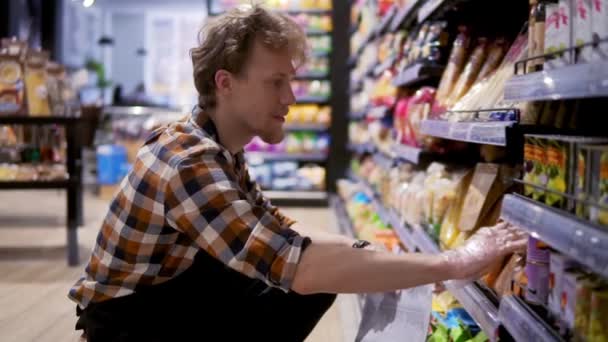 Kaufmann im Supermarkt sortiert Artikel in einem Regal, notiert das Fehlen von Produkten, sortiert Artikel in einem niedrigeren Regal — Stockvideo