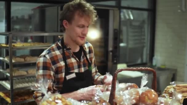 Commis de magasin positif et souriant en tablier noir mettant de la pâtisserie fraîche, des muffins sur la vitrine debout dans le beau magasin avec des produits de boulangerie. Vue de face — Video