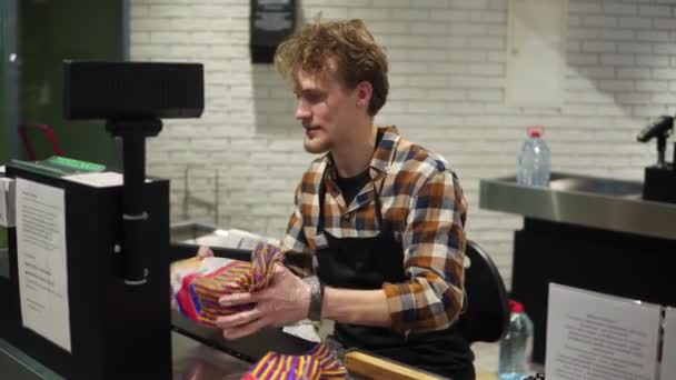 Belle caissière jeune homme barbu sert le client, produits de caisse sur le comptoir. Travailleur positif, souriant — Video