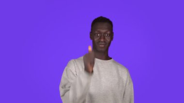 Beyaz kazaklı, gülümseyen, Afro-Amerikalı genç bir adam stüdyoda mavi duvarın ardından izole edilmiş bir jest yapıyor.