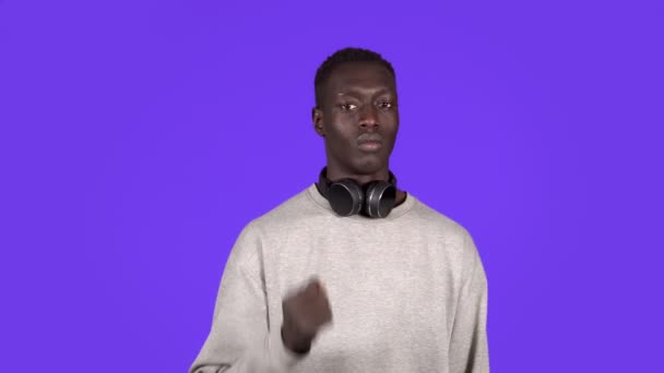 青い背景のスタジオに隔離されたポーズの白いシャツの若いアフリカ系アメリカ人男性の肖像画。人々の感情ライフスタイルの概念。手でストップジェスチャーを表示,首にヘッドフォン — ストック動画