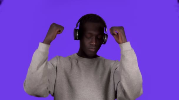 Homem africano agradável em camisa branca e fones de ouvido dançando relaxante com os olhos fechados sobre fundo azul. Movimento lento — Vídeo de Stock