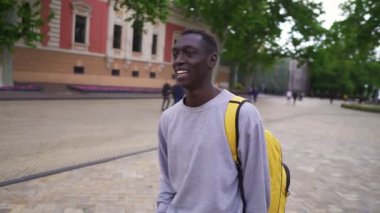 Pozitif, gülümseyen Afro-Amerikan bir adam şehirde sarı sırt çantasıyla yürüyor, gündelik giysiler giyiyor. Etrafına bak, eski sokakların geçmişi. Yavaş çekim