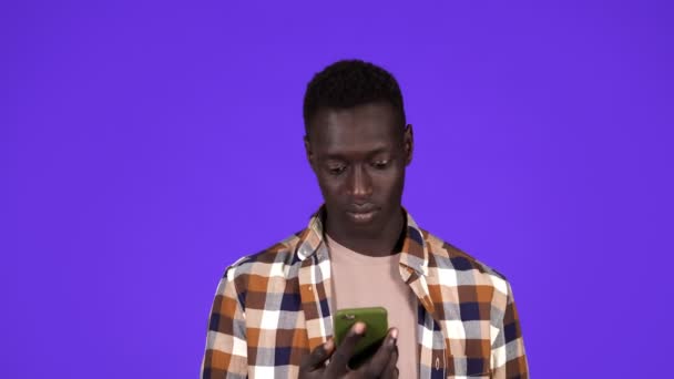 Молодой черный мужчина держит телефон скрещивая пальцы за удачу, смотрит на мобильный экран и врывается улыбаясь, победитель. Изолированный на синем фоне — стоковое видео