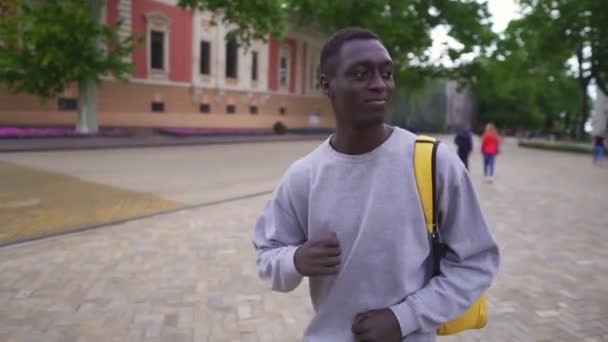 Jeune homme noir drôle portant des vêtements décontractés, sac à dos jaune profitant de la danse de marche de plaisir sur le trottoir par la rue de la ville. Mouvement lent — Video