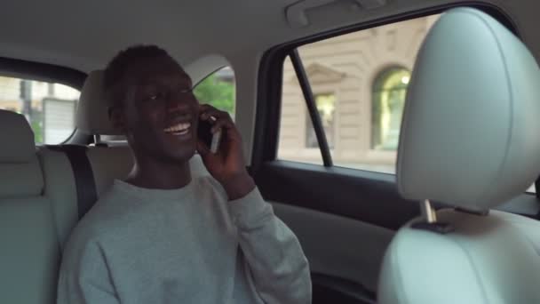 Hombre africano positivo y sonriente con ropa casual que habla por teléfono celular mientras monta en el asiento trasero de la cabina de taxi. alegre negro pasajero chico contestar llamada teléfono, hablando con amigos — Vídeo de stock