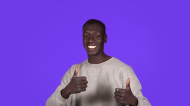 Genç Afro-Amerikalı adam gülümsüyor ve mavi arka planda mutlu bir şekilde dans ediyor. Durdurulamaz eğlence, mutluluk, baş parmaklar, insani duygular