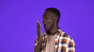 Kısa saçlı Afro-Amerikan bir adam esniyor, yüzüne dokunuyor, yorgun ve uykulu bir şekilde stüdyoda mavi duvarın arkasında dikiliyor. Yan görünüm