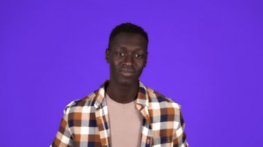Mutlu, gülümseyen Afrikalı Amerikalı adam mavi arka planda iyi bir ruh hali içinde. Baş parmaklarıyla hareket ediyor, jest yapıyor, ekose gömleğine dokunuyor - insani duygular.