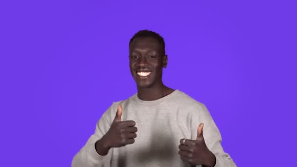 Genç Afro-Amerikalı adam gülümsüyor ve mavi arka planda mutlu bir şekilde dans ediyor. Durdurulamaz eğlence, mutluluk, baş parmaklar, insani duygular — Stok video